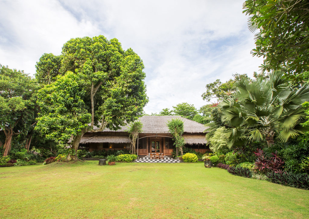 Casa de Nipa, a grand scale nipa hut with thatched roof surrounded by tropical vegetation.