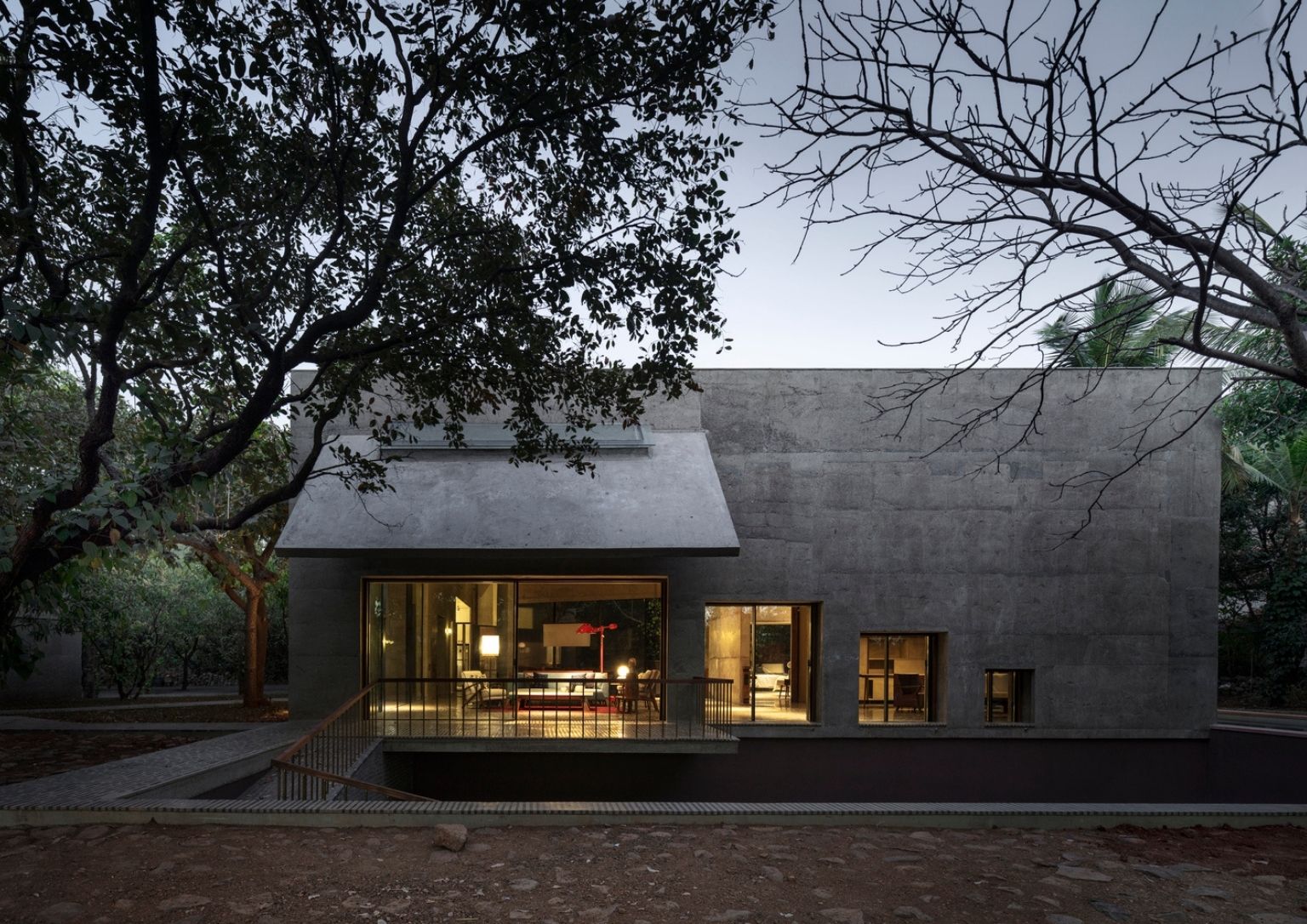 A minimalist concrete home lit from inside with warm yellow lights surrounded by trees.