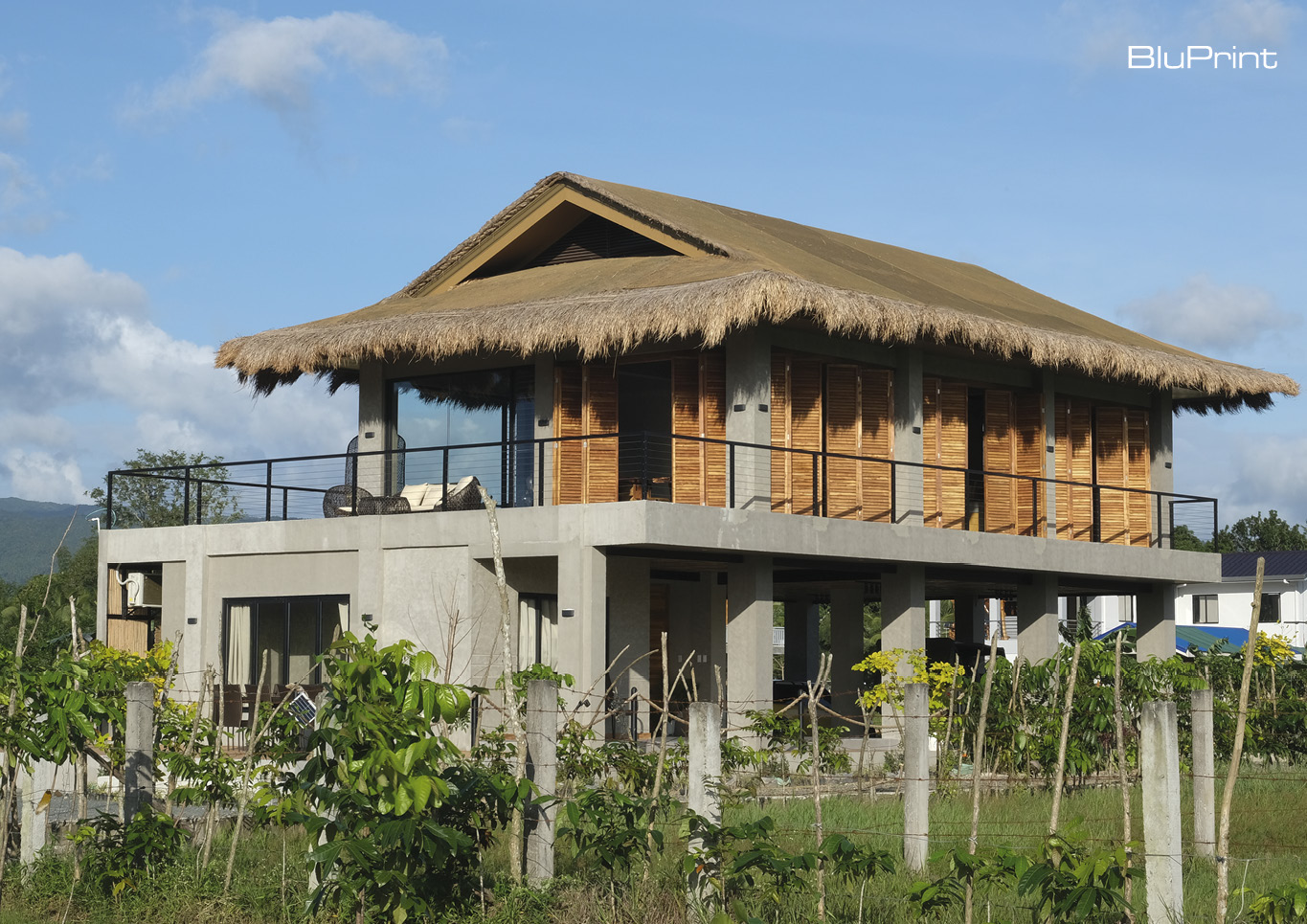 A modern bahay kubo design on concrete stilts and thatched roof in the middle of a field.