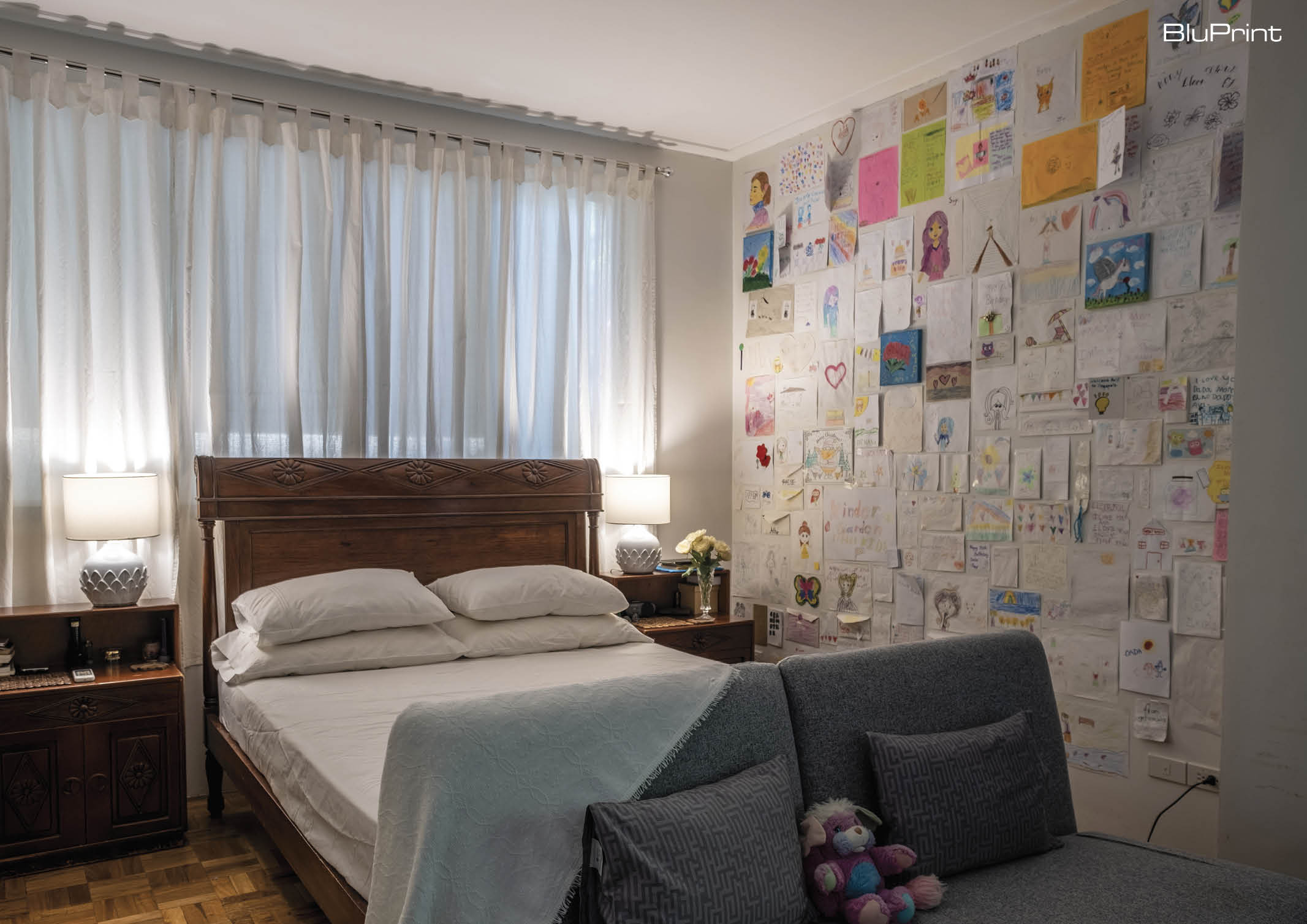 A kid's bedroom with a wooden bed frame and side tables. A two-seater couch is at the foot of the bed, and a wall displays the child's art work