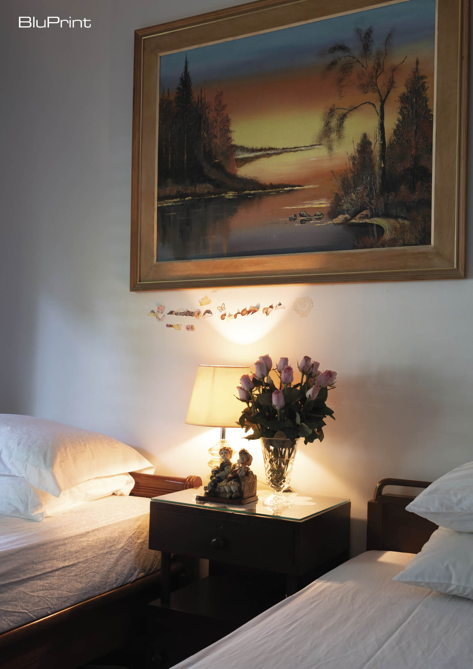 A bedroom with two twin beds and a side table with a floral arrangement in between. An oil painting hangs above it.