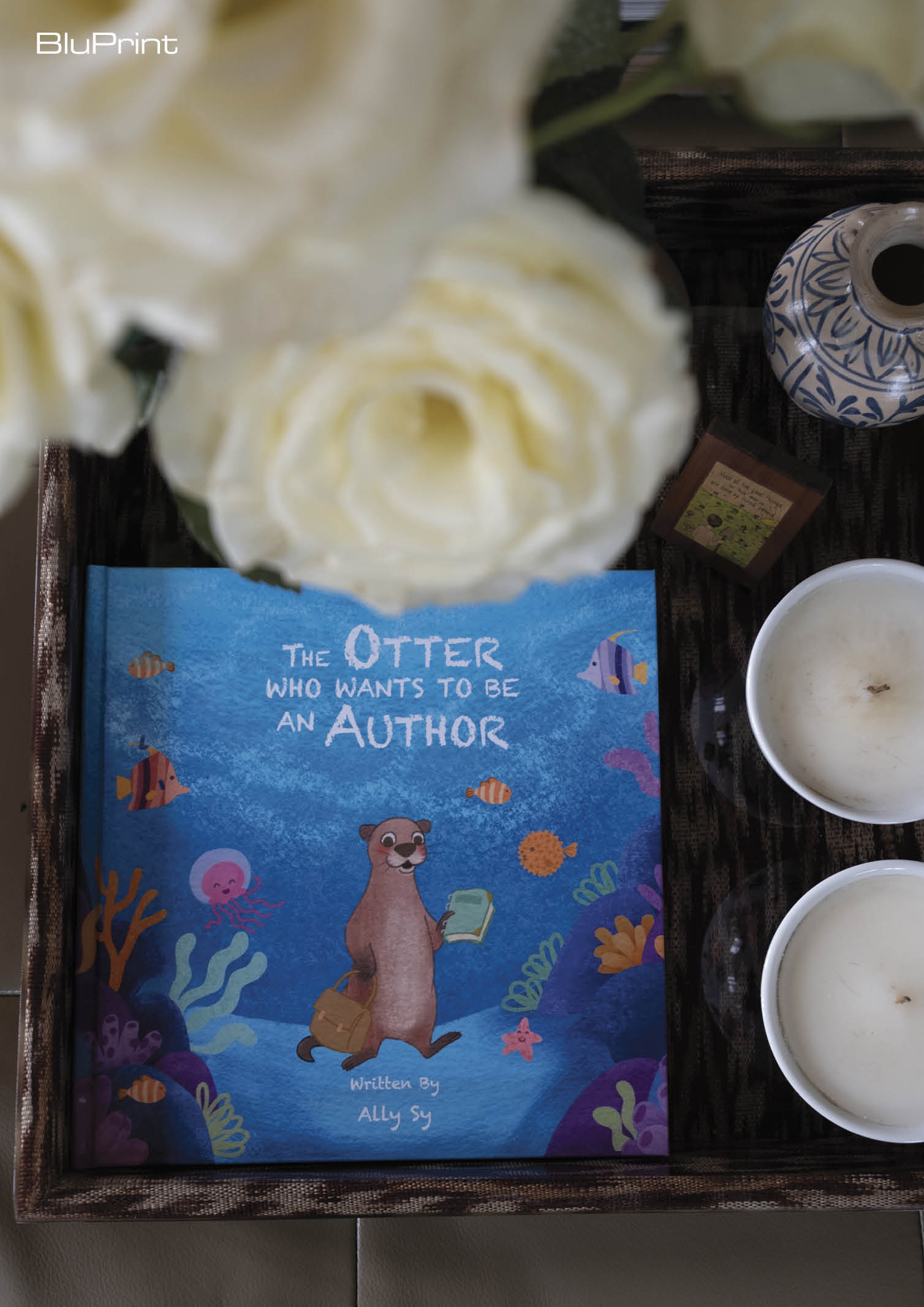 Image of a book cover of the "The Otter Who Wants to Be an Author" next to candles and a floral arrangement
