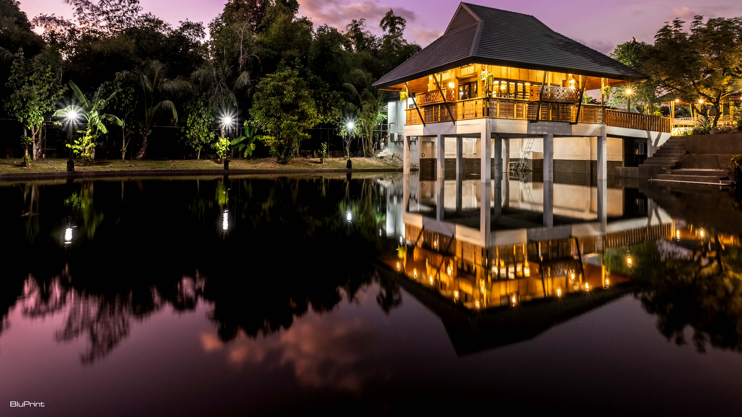 A night time shot of a modern bahay kubo lit up from inside, the lights reflecting on the small lake it sits in.