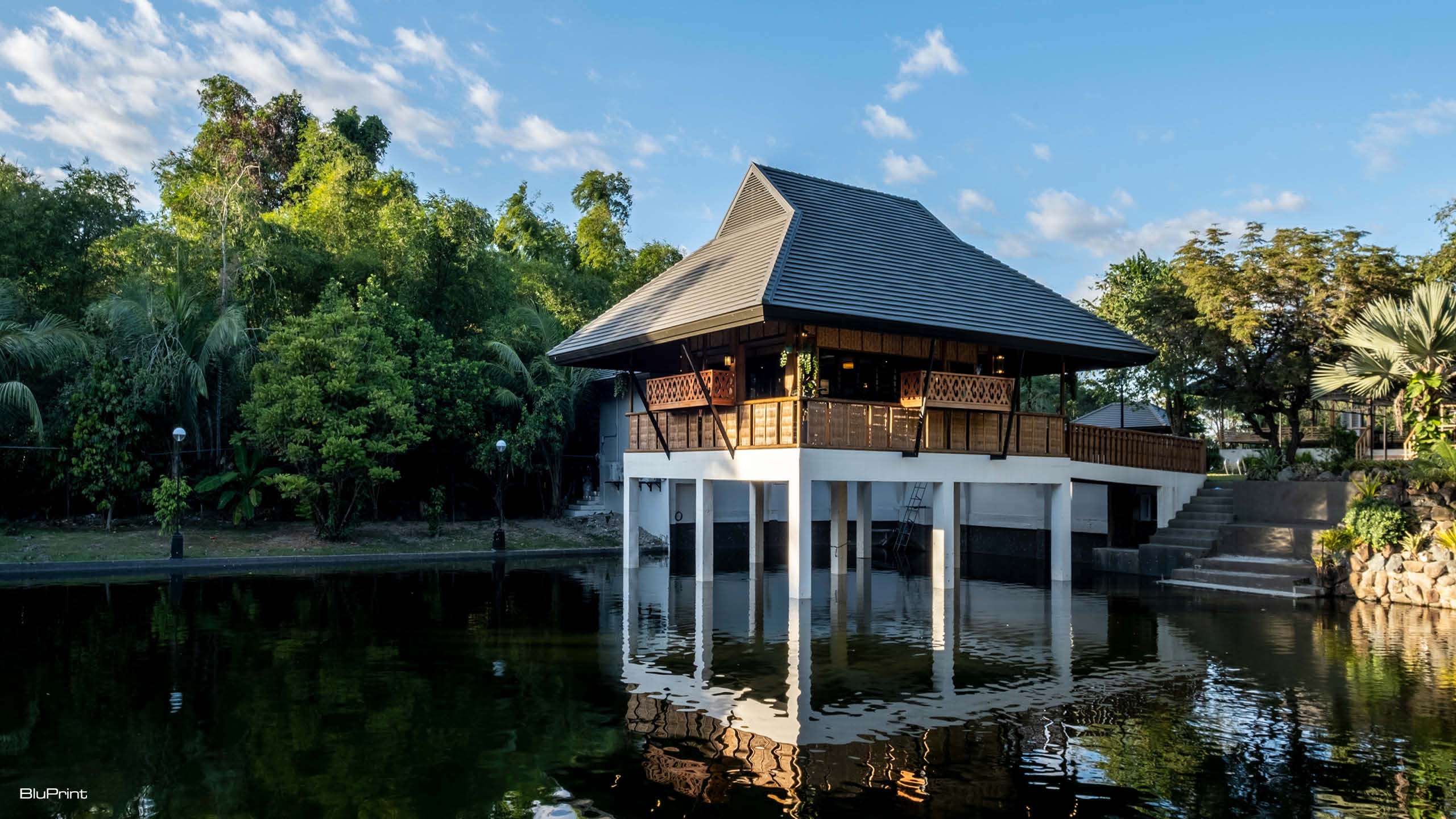A modern bahay kubo with a pitched roof sitting above a small lake on concrete stilts.