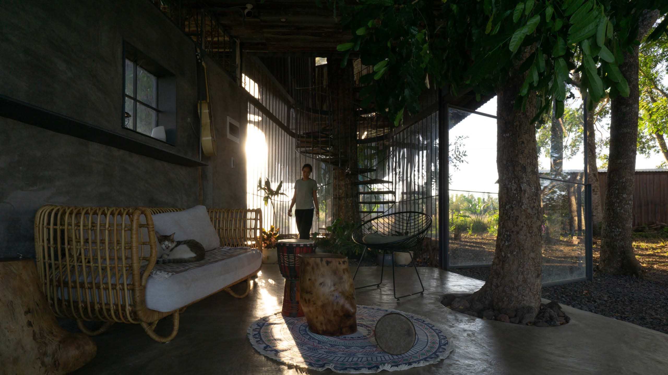 A woman next to a wooden spiral staircase walks into an open living room surrounded by nature.