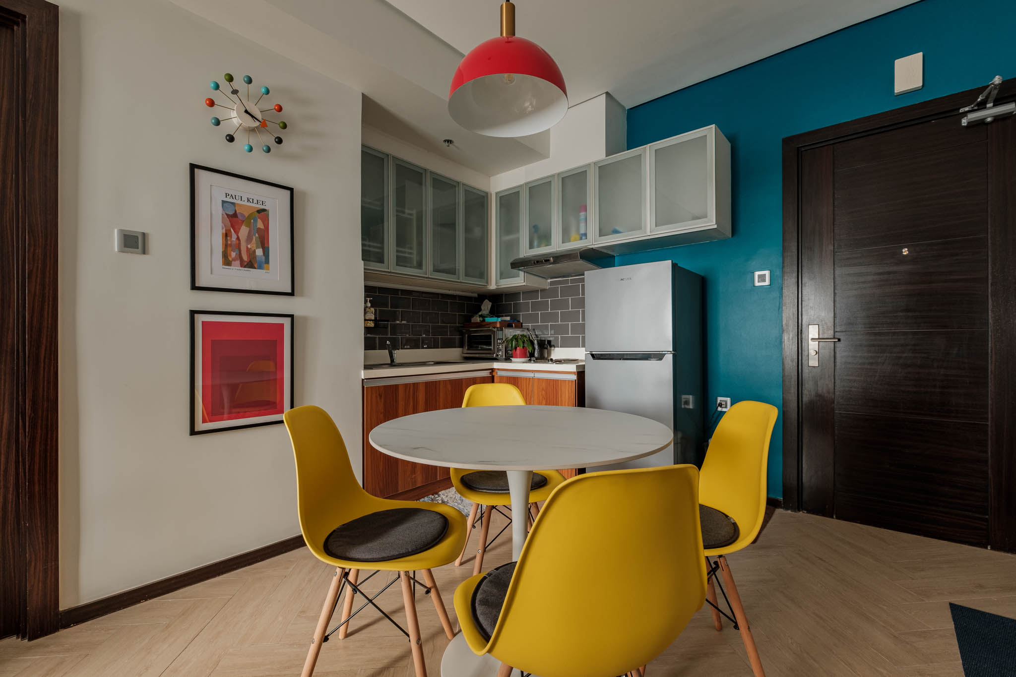 Modern apartment interior with teal accent wall, red pendant lamp, and yellow chairs beside a round dining table.