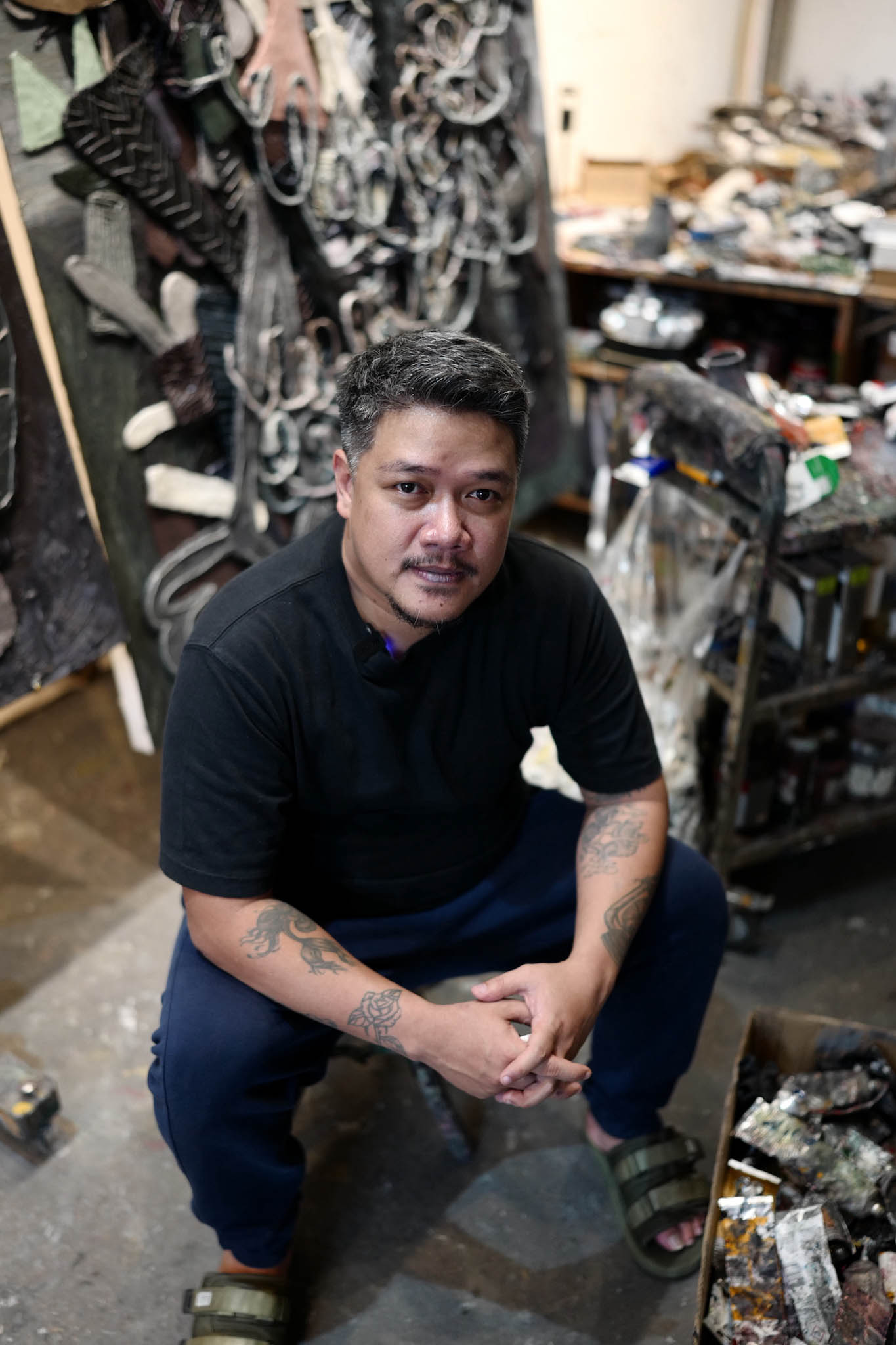 An artist squats in his studio surrounded by his art materials.