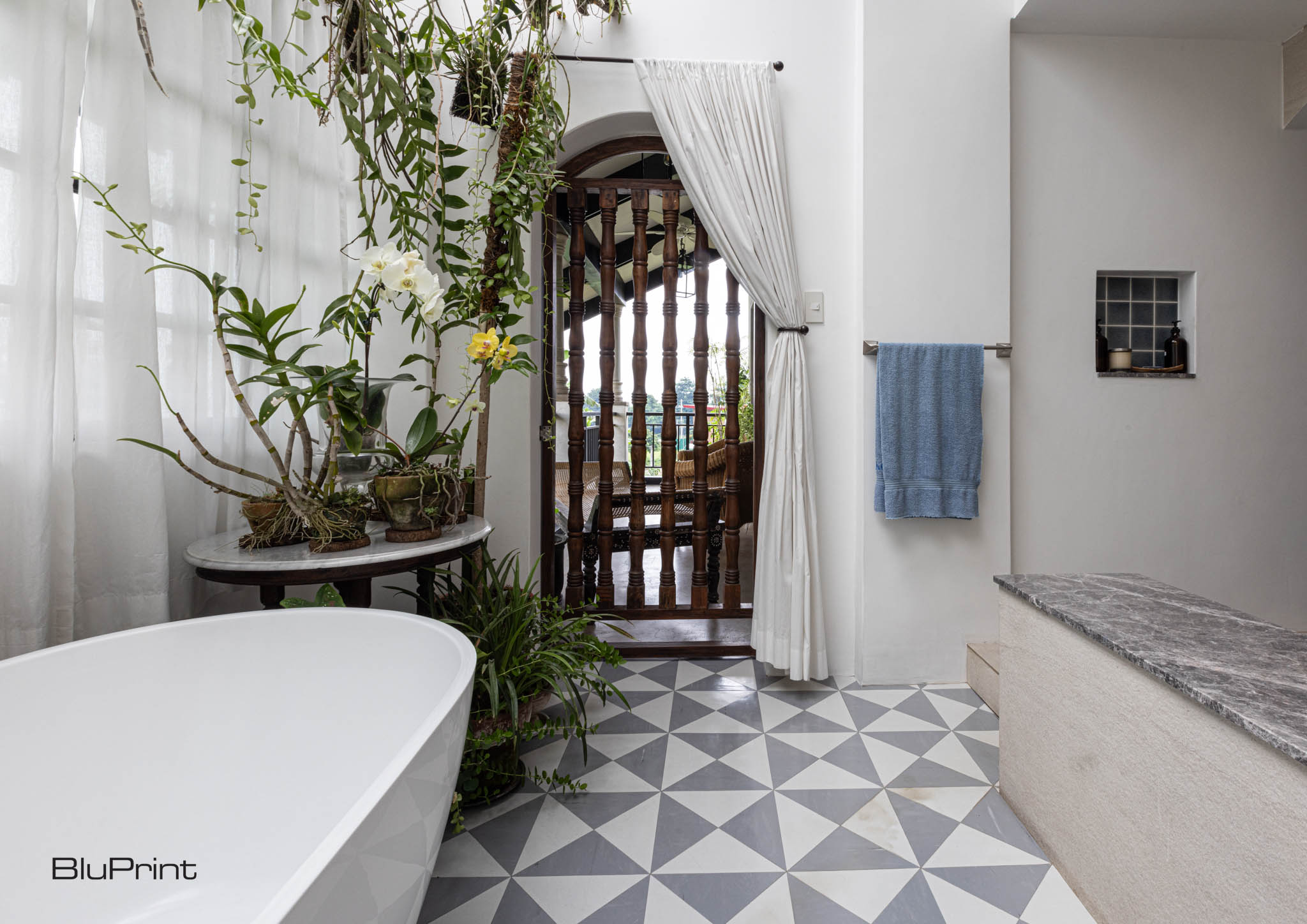 A large bathroom filled with plants, with a large white tub, patterned tile, fabric curtains.