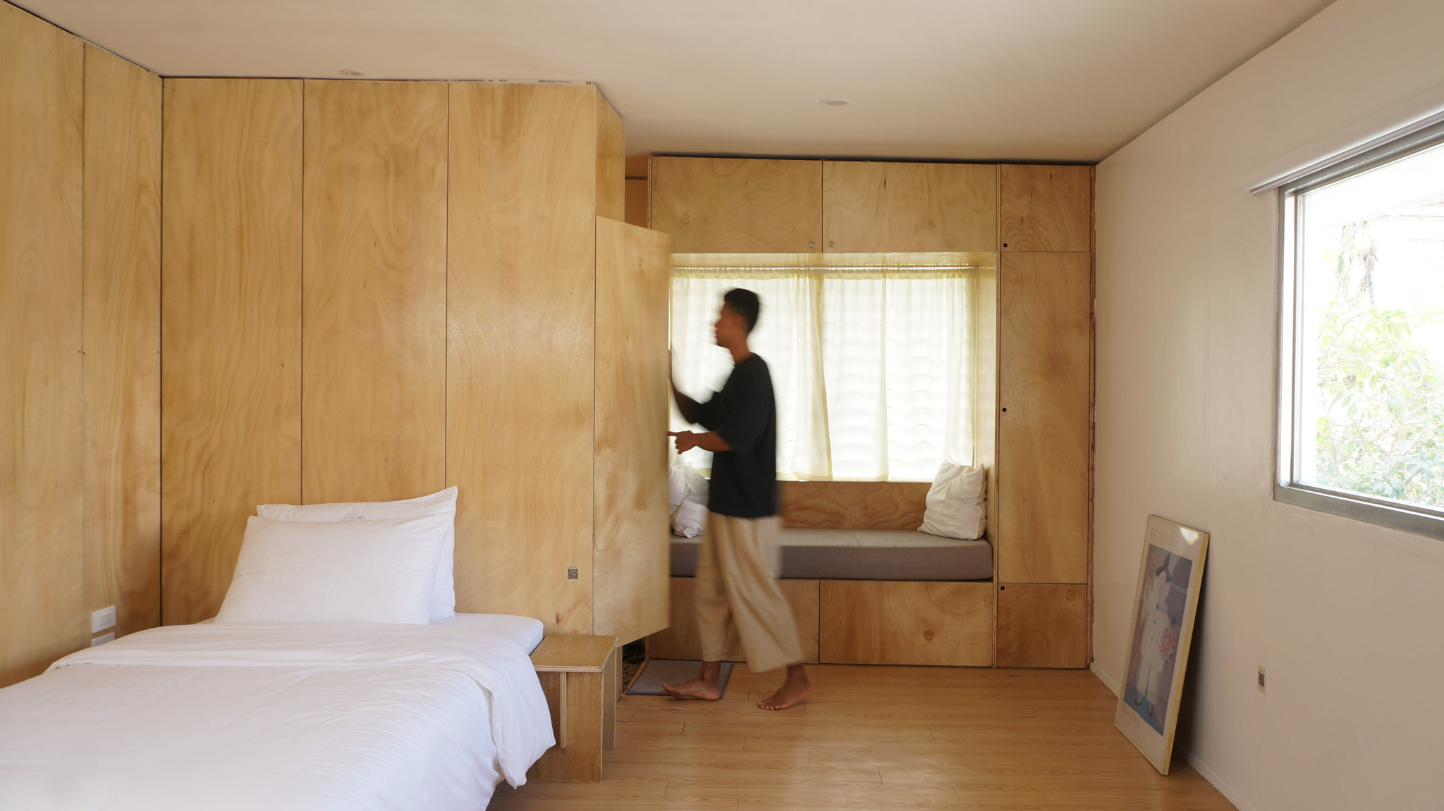 A man opens a closet next to a window seat surrounded by built-in cabinetry. A bed with white linens can also be seen.