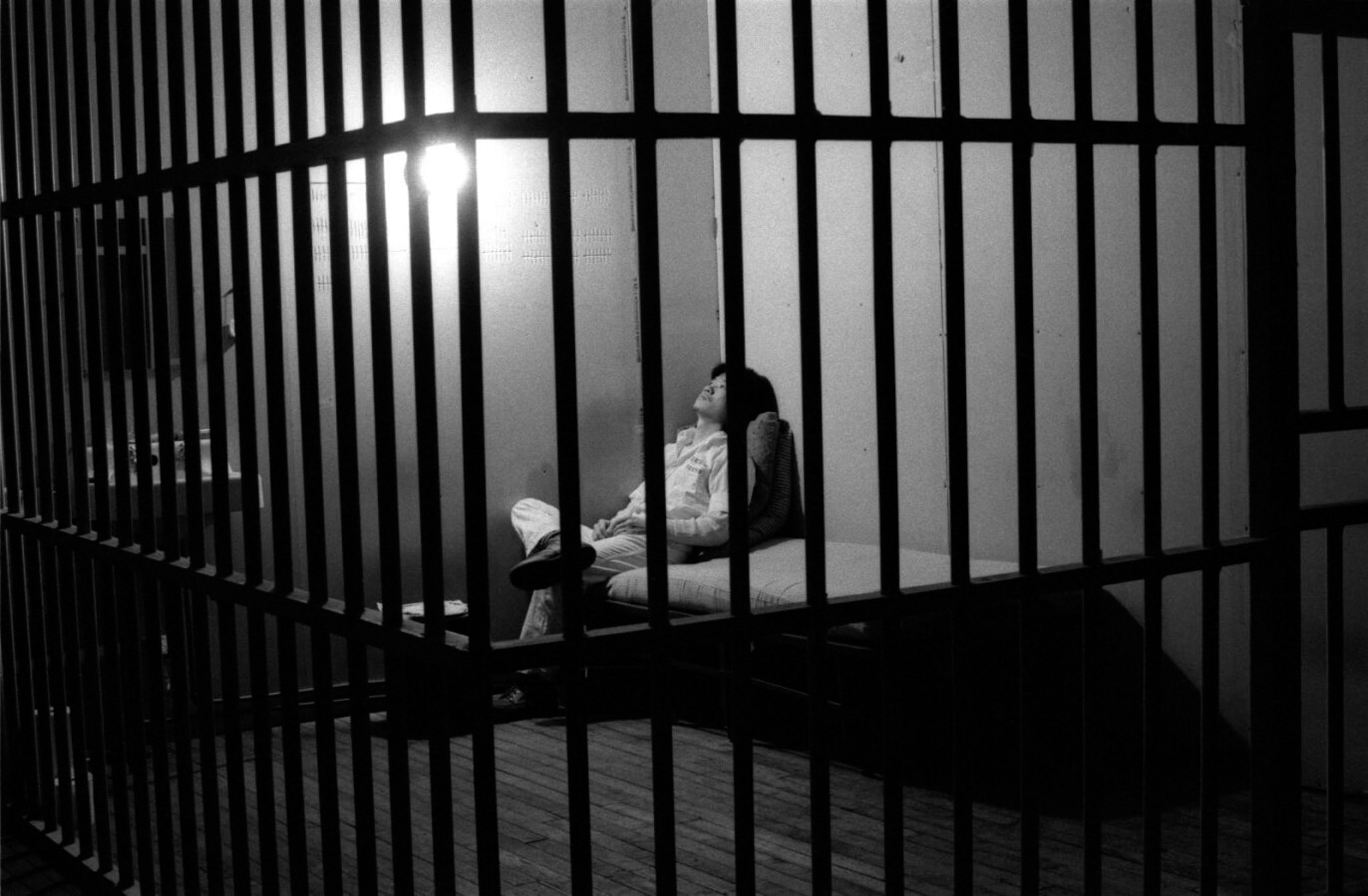 A black and white photograph of a person sitting in a jail cell. 