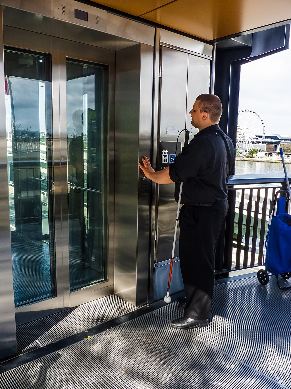 A person with visual-impairment using braille and tactile flooring to locate an elevator. Photo by John Robert McPherson. Source: Wikimedia Commons.