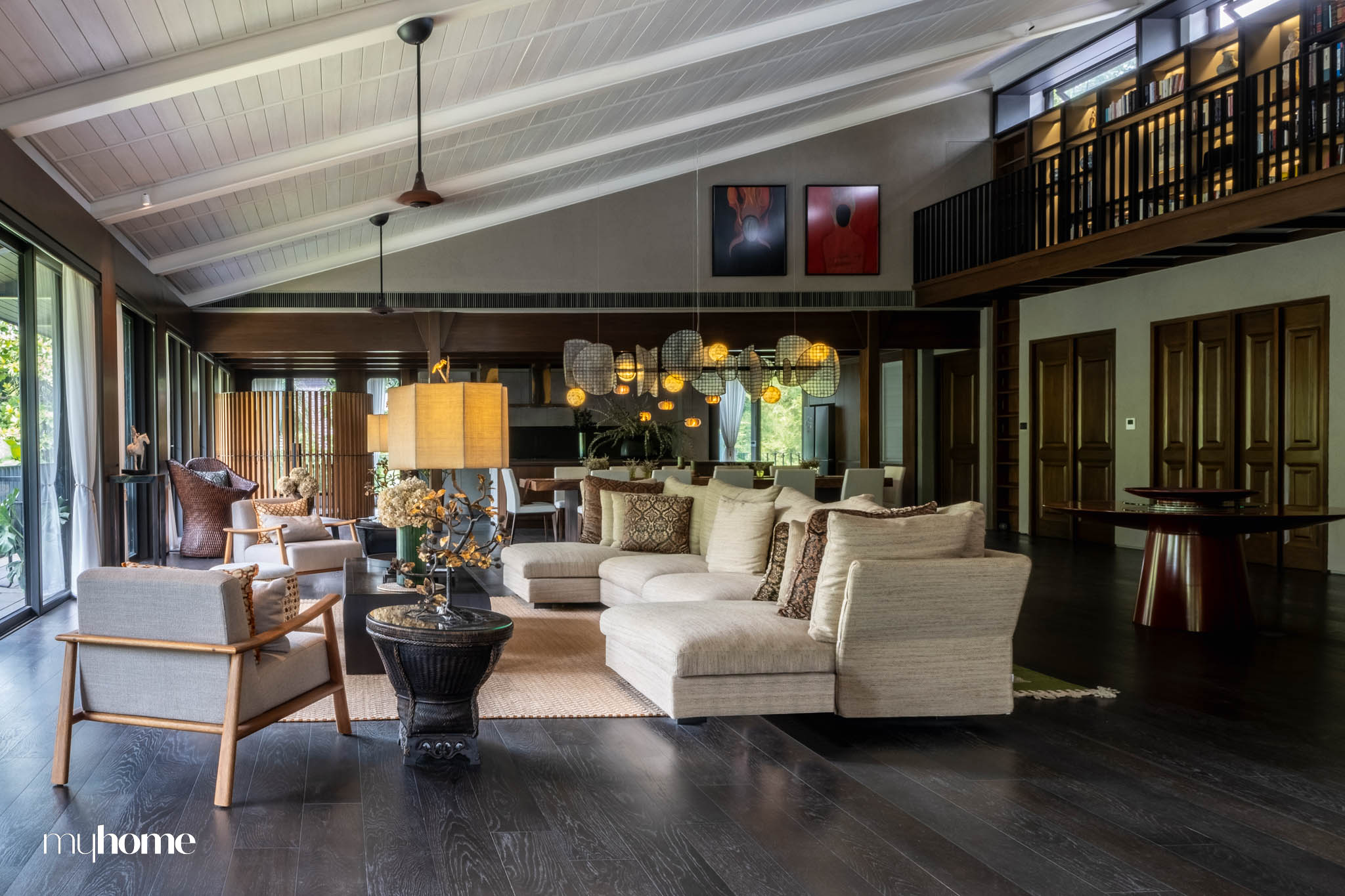 A spacious living area in Instia House, showcasing a harmonious blend of traditional Filipino design with modern elements. The room features wooden furniture, woven textures, and a large sectional sofa, all set against dark wood flooring and a vaulted white ceiling. Large windows allow natural light to fill the space, highlighting the fusion of classic and contemporary aesthetics.