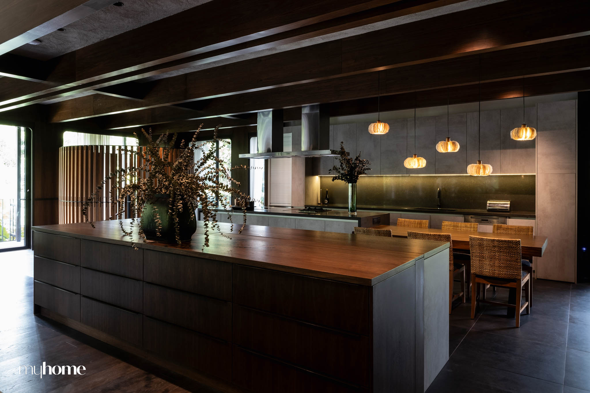 A modern kitchen interior at Instia House featuring a blend of Filipino and contemporary design. The space includes a large wooden island, woven dining chairs, and pendant lighting, with dark wood tones and natural materials that reflect Filipino craftsmanship.