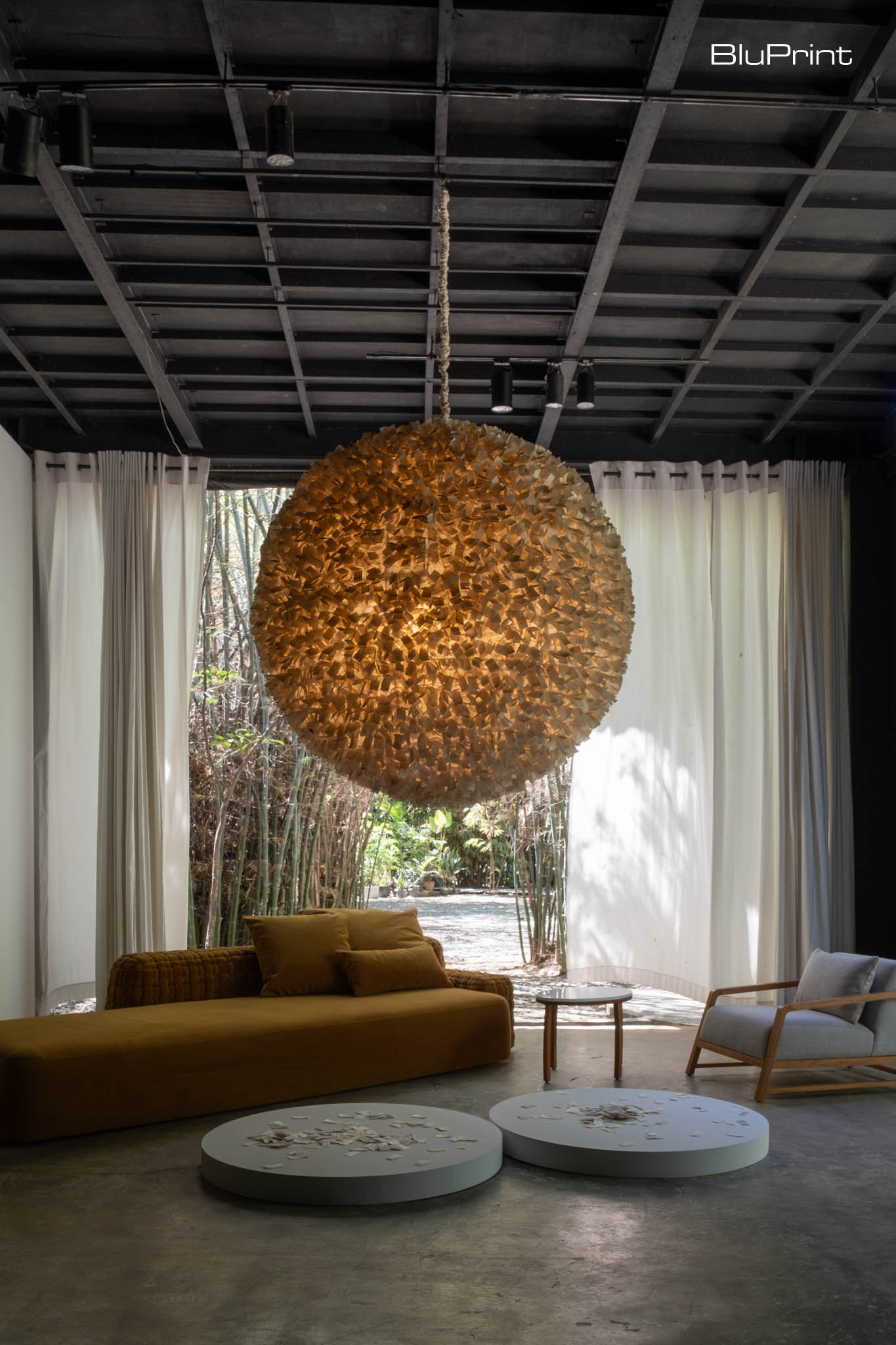 A large, spherical pendant light, textured with layered materials by OMO Furniture hangs from the ceiling, casting a warm, ambient glow over the space. Below it, a minimalist, mustard-colored daybed pairs with a matching cushion, positioned beside two circular coffee tables. A simple wooden armchair with light upholstery completes the seating arrangement, adding to the room's earthy and contemporary aesthetic.